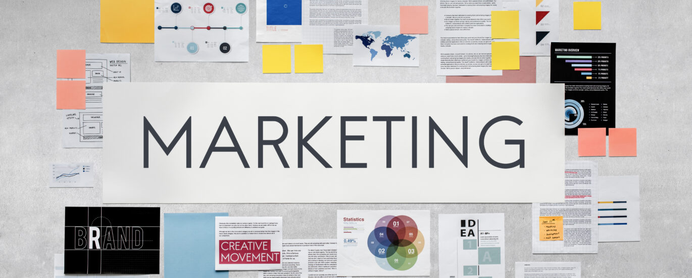 Overhead view of a diverse array of marketing strategy materials spread out on a gray surface, featuring diagrams, graphs, sticky notes, and a large 'MARKETING' banner in the center, suitable for a discussion on best practices for adding French subtitles to marketing videos.