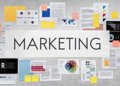 Overhead view of a diverse array of marketing strategy materials spread out on a gray surface, featuring diagrams, graphs, sticky notes, and a large 'MARKETING' banner in the center, suitable for a discussion on best practices for adding French subtitles to marketing videos.