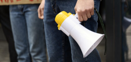 A person holds a yellow megaphone at a protest with a partially visible sign reading 'WAKE UP' in the background, illustrating the significance of inclusive communication such as Spanish subtitles in public announcements and emergency alerts.